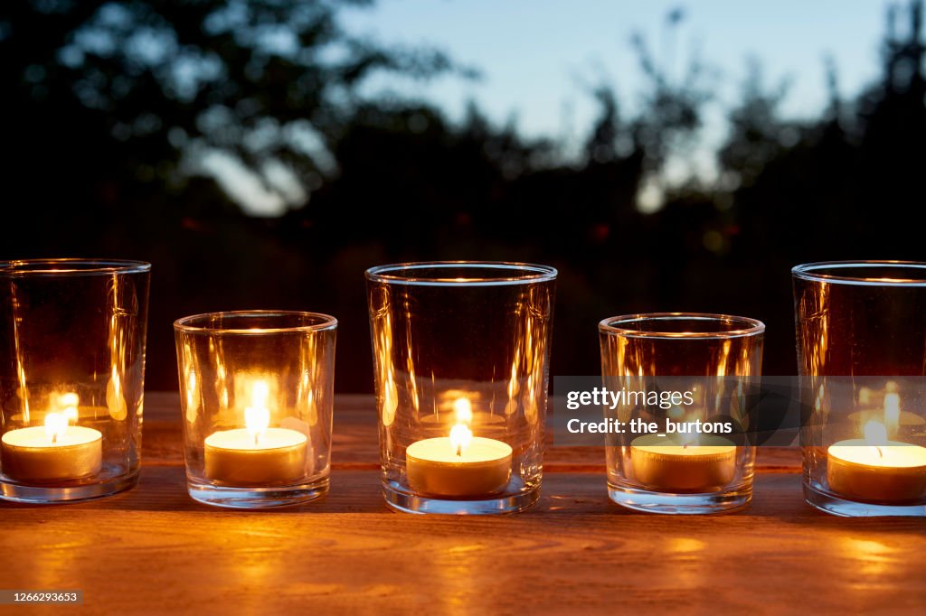 Lit tea light candles in glasses in a row on table in garden during sunset