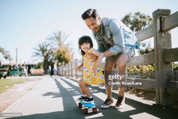 father helps young daughter ride skateboard - happiness imagens e fotografias de stock