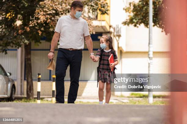 lovely school girl holding her father's hand as they walk to school, both wearing masks - parent daughter school uniform stock pictures, royalty-free photos & images