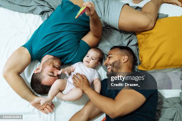 smiling gay couple playing with adopted baby girl on bed - gay person stock pictures, royalty-free photos & images