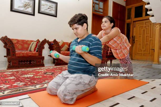 a boy doing biceps dumbbell exercise and a girl pushing him from back in side the domestic room on exercise mat. - pakistani boys stockfoto's en -beelden