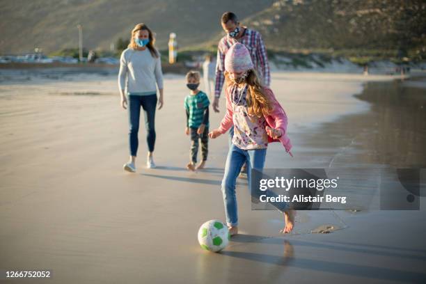 girl wearing mask kicking soccer ball on beach walk with family - football face mask stock pictures, royalty-free photos & images