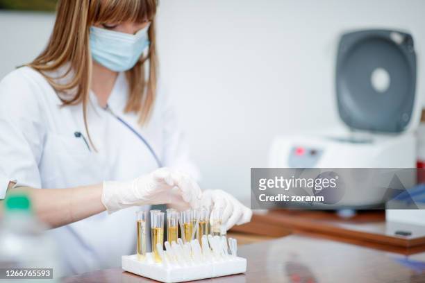 mujer usando máscara facial y guantes poniendo tubos con muestra de orina en el estante - urine sample fotografías e imágenes de stock