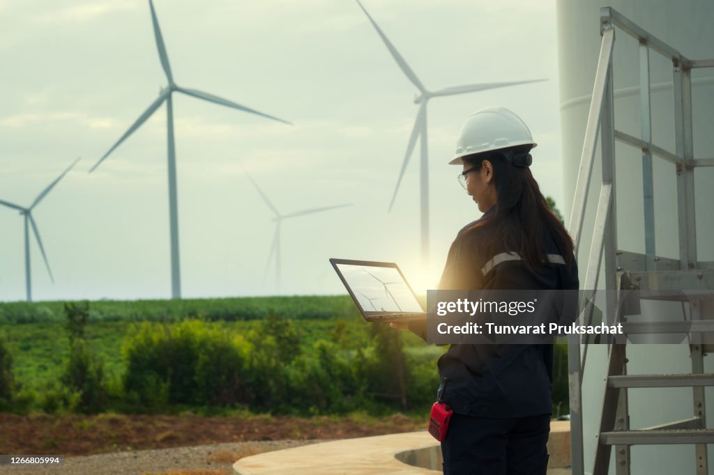 Female engineer wearing hard hat standing with digital tablet against wind turbine.