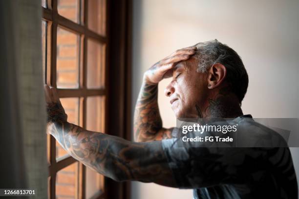 hombre maduro preocupado de pie en casa, cerca de una ventana, con la mano en la cabeza - only mature men fotografías e imágenes de stock