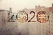 hahdmade painted numbers 2020 with sad face with closed eyes in protective medical mask on splashed by rain foggy glass on blue window, concept photo self-isolation, coronavirus, covid - 19