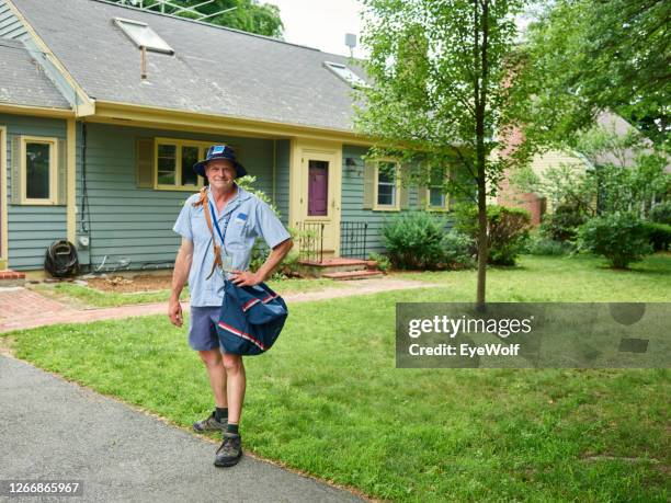 a mail man walking away from a house with a mail sack hanging off his shoulder, looking into camera smiling. - postbode stockfoto's en -beelden
