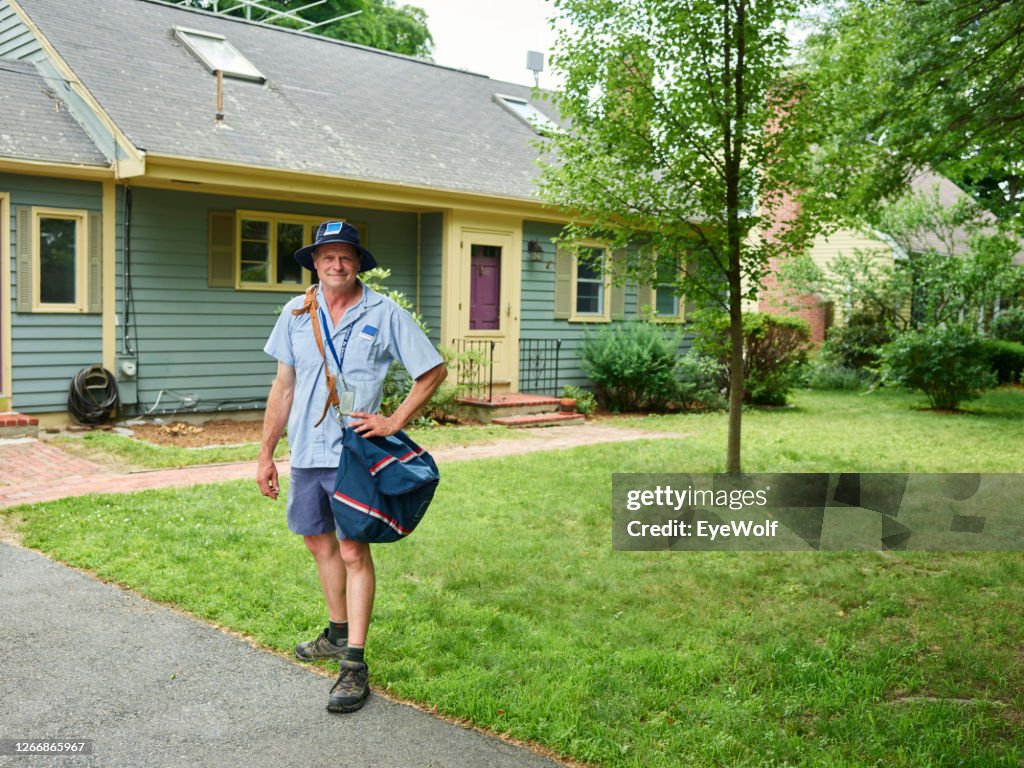 A mail man walking away from a house with a mail sack hanging off his shoulder, looking into camera smiling.