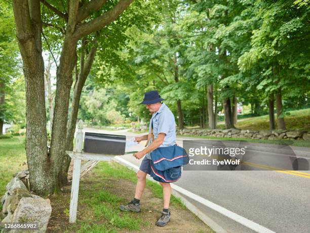 a mailman delivering a letter into a mailbox on the side of the road in the summer. - postbode stockfoto's en -beelden