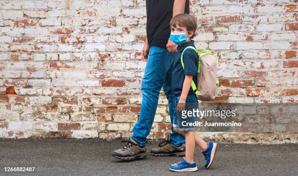 father holding his son hand walking him back to school, boy is wearing protective face mask and carry a backpack in his shoulders - protective face mask side stock pictures, royalty-free photos & images