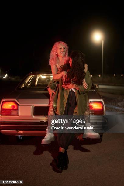 two girl friends leaning on car having fun at night - auto daten stock pictures, royalty-free photos & images