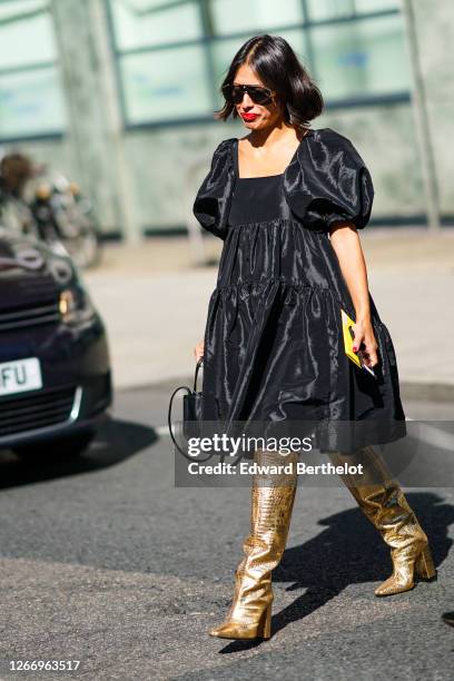 Guest wears sunglasses, a black shiny pleated and gathered dress with puff shoulders, golden shiny pointy high heeled long boots, during London...