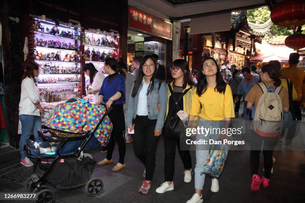 Three Chinese girls, tourists, walk in a narrow alley in the historical Yuyuan quarter, shops are seen on either side, a mother with a baby stroller...