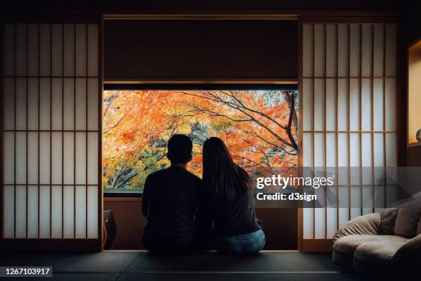 a loving young asian couple sitting side by side by the window in a traditional japanese style apartment, chatting and enjoying the beautiful nature autumn scenics during the day - japanese culture foto e immagini stock