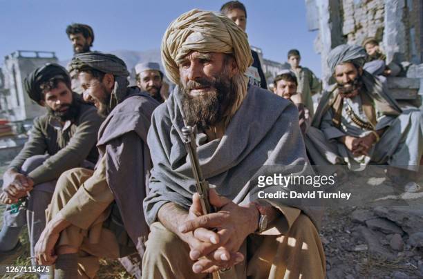 An Afghan man wearing a lungee turban and patoo shawl, the barrel of his rifle held between his hands, with similarly dressed men in the background,...