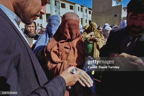 Afghan women, some dressed in niqabs, others in hijabs, with one woman holding her hand out to be marked by a man holding a pen at an registration...