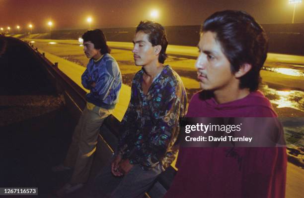 Three young Mexican men sit on a wall above a flood control channel as night falls on the border between Mexico and California in the United States,...