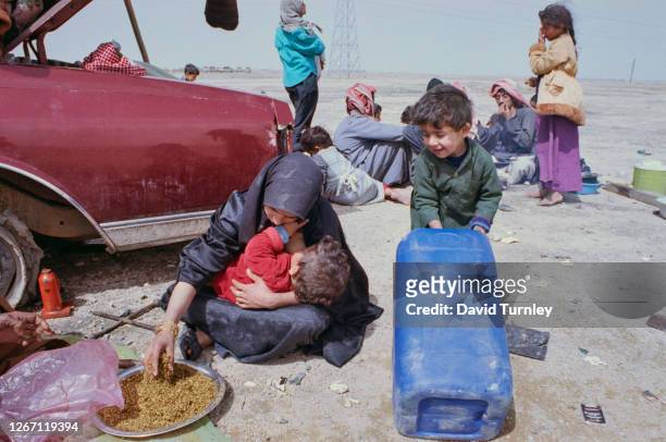 Woman wearing a black hijab feeds her child from a plate of food before her, as she cradles the child in her arms, as a small boy pulls a large...