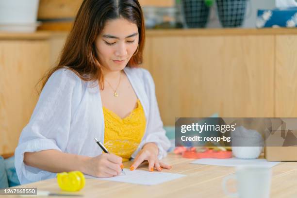 young woman writing letter to put inside gift box in living room at home - grant writer stock pictures, royalty-free photos & images