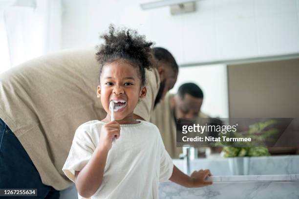 african cute kid brushing her teeth by toothbrush while her father standing behind in the bathroom. dental hygiene, healthcare concept. - tooth bonding stock pictures, royalty-free photos & images
