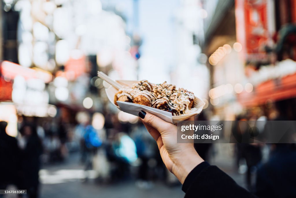 Personal perspective of a female traveller holding freshly made traditional Japanese street-style snack takoyaki (octopus balls) against downtown city street while visiting Osaka