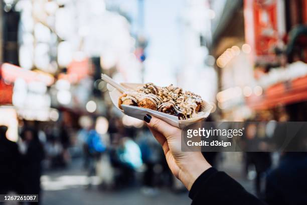 personal perspective of a female traveller holding freshly made traditional japanese street-style snack takoyaki (octopus balls) against downtown city street while visiting osaka - osaka prefecture stock pictures, royalty-free photos & images