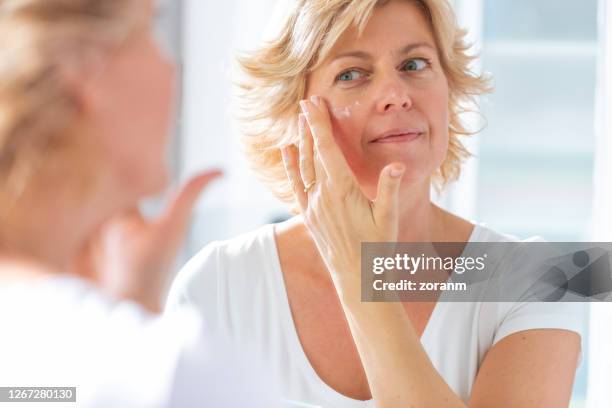 medio volwassen vrouw die spiegel bekijkt en moisturizer op haar gezicht uitspreidt - gezichtscrème stockfoto's en -beelden