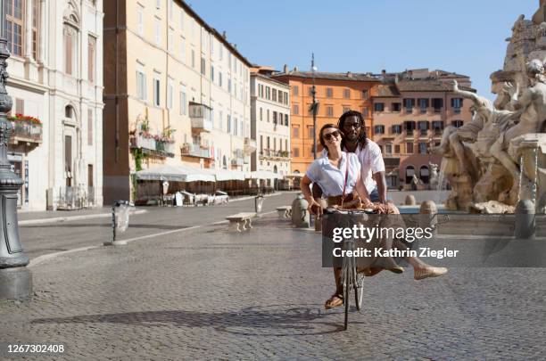 beautiful biracial couple riding bicycle in deserted piazza navona, rome, italy - rome italie photos et images de collection