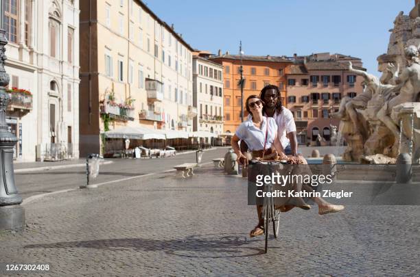 beautiful biracial couple riding bicycle in deserted piazza navona, rome, italy - rome italië stockfoto's en -beelden