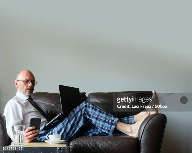 businessman working from home on couch in dress shirt and pyjama pants with laptop and mobile smartphone, stuttgart, germany - working from home stock-fotos und bilder