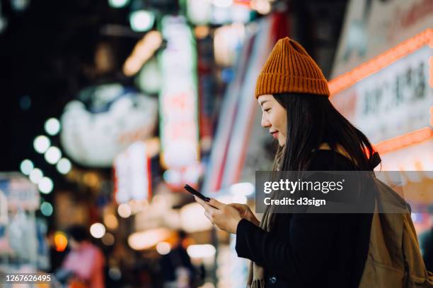 stylish young asian female traveller with backpack using mobile app device on smartphone to navigate location while exploring in busy downtown city street at night - shinsekai osaka foto e immagini stock