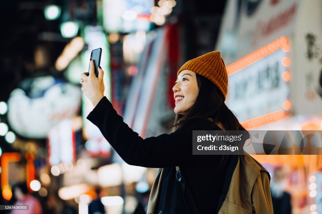 Cheerful and stylish young Asian female traveller with backpack taking a selfie with smartphone while exploring and strolling in busy downtown city street at night in Osaka, Japan