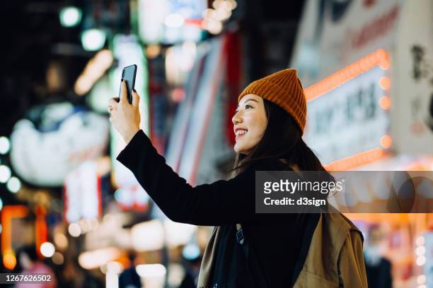 cheerful and stylish young asian female traveller with backpack taking a selfie with smartphone while exploring and strolling in busy downtown city street at night in osaka, japan - osaka city 個照片及圖片檔