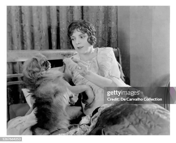 Silent film Actress Alma Rubens in bed giving a treat to a Pekingese dog in an unknown film from the 1920’s, United States.