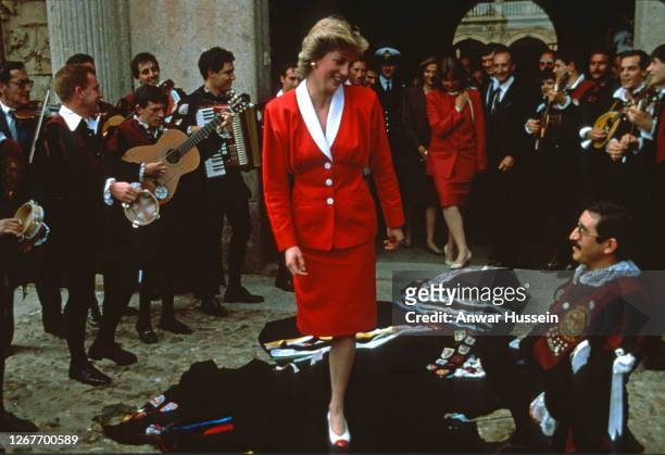Diana, Princess of Wales, wearing a red skirt suit with a white collar designed by Bruce Oldfield, walks over musician's cloaks which have been...