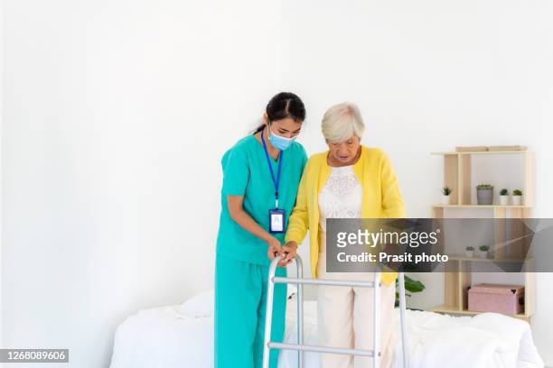 smiling asian nurse assisting senior woman to get up from bed. caring nurse supporting patient while getting up from bed and move towards walker at home. helping elderly disabled woman standing up. - doctor on the move stock pictures, royalty-free photos & images
