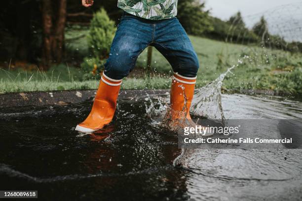 child wearing orange wellies jumping in a deep puddle - oranger stiefel stock-fotos und bilder