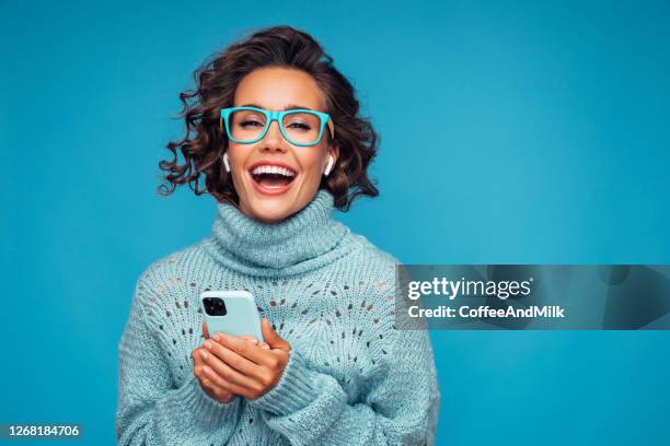 スマートフォンで青い背景の前に立っている美しい女性 - happy glasses ストックフォトと画像