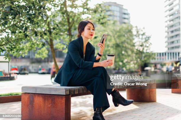 young businesswoman using smart phone on bench at park, with coffee cup on hand - stylish woman streets europe cellphone stockfoto's en -beelden