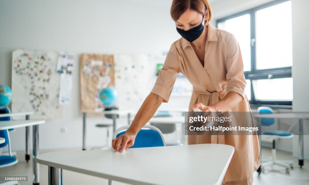 Woman teacher with face mask disinfecting desks, back to school concept.