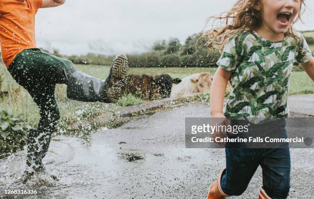 two children in welly boots play in a huge puddle - children only stock-fotos und bilder