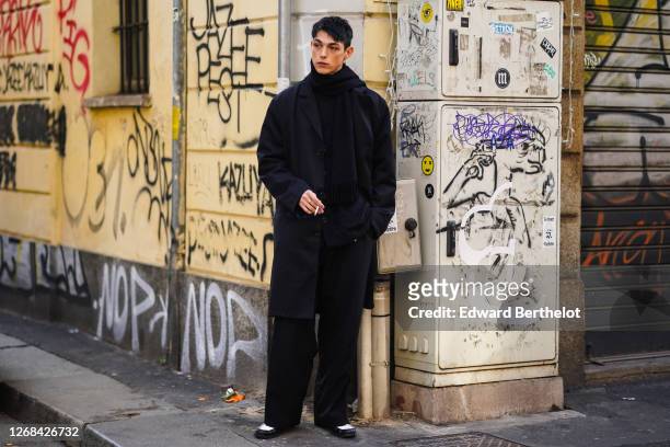 Model wears a black pullover, a scarf, a black oversized long coat, flared pants, shoes, outside Magliano, during Milan Fashion Week Menswear...