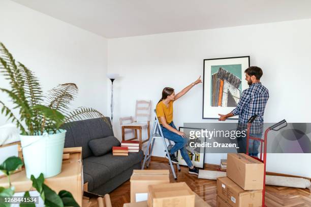 pareja en movimiento en nuevo apartamento, mujer y hombre poner imagen en la pared - decorating fotografías e imágenes de stock