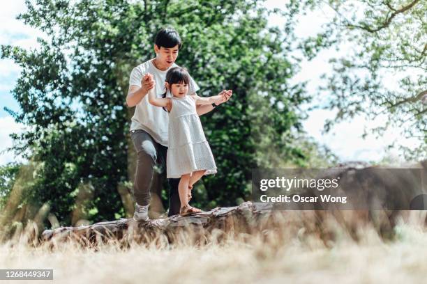 caring young asian father holding hands of his little daughter and assisting her to walk along a tree trunk outdoor on a sunny day - china balance stock-fotos und bilder