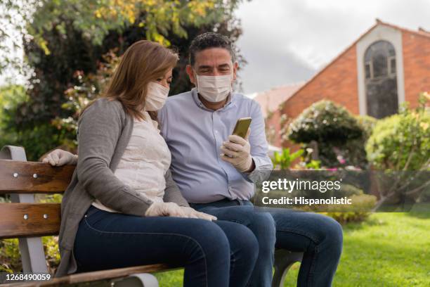 happy couple wearing a facemask outdoors while looking at social media on a cell phone - flatten the curve icon stock pictures, royalty-free photos & images