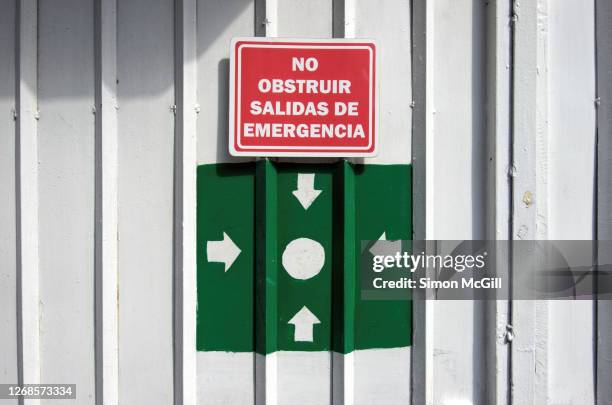 metal gate with spanish-language warning sign: 'no obstruir. salidas de emergencia' (do not obstruct. emergency exits) and a hand-painted evacuation assembly point sign - obstruir stock pictures, royalty-free photos & images