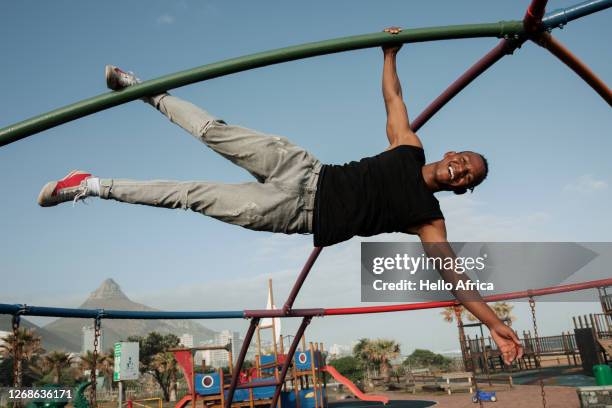 handsome strong young man hanging by an arm and a leg from a climbing structure - monkey bars fotografías e imágenes de stock