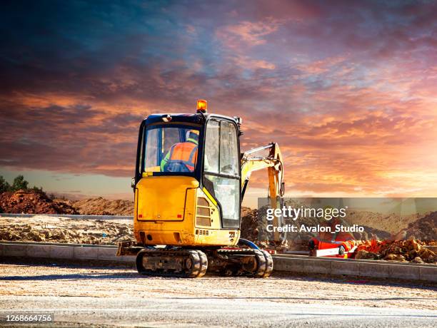 mini excavator and worker at a construction site against the setting sun - construction vehicle stock pictures, royalty-free photos & images