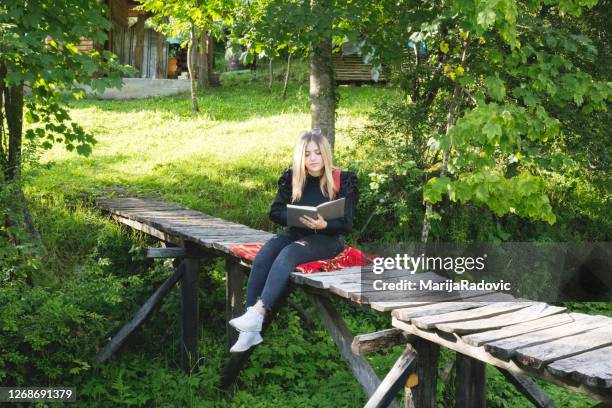 junge blonde reisende frau lesen buch auf brücke im wald. grüner hintergrund - roman bridge stock-fotos und bilder