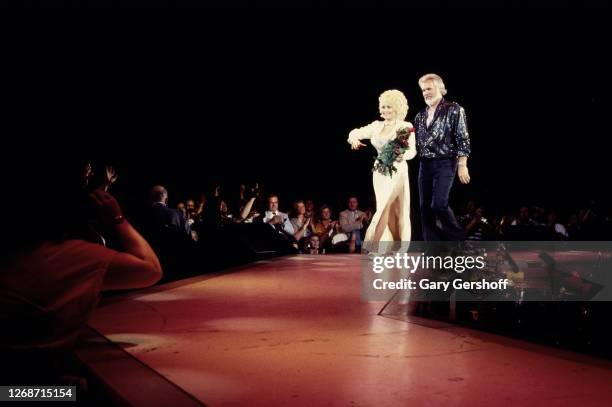 American Country musicians Dolly Parton, holding a bouquet of red roses, and Kenny Rogers walk onstage during a performance at Nassau Coliseum,...